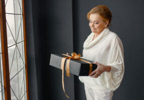 Seniour woman has a gift box. Woman standing near big window and posing for a photo. Red-haired woman wearing white clothes.