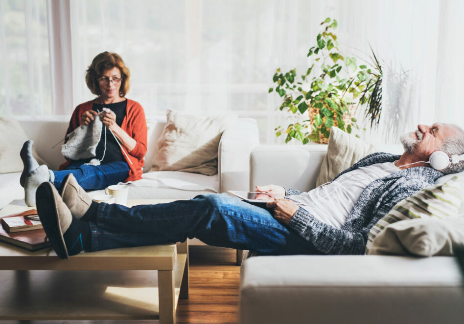 Happy senior couple relaxing at home. A woman knitting and a man with tablet listening to music.